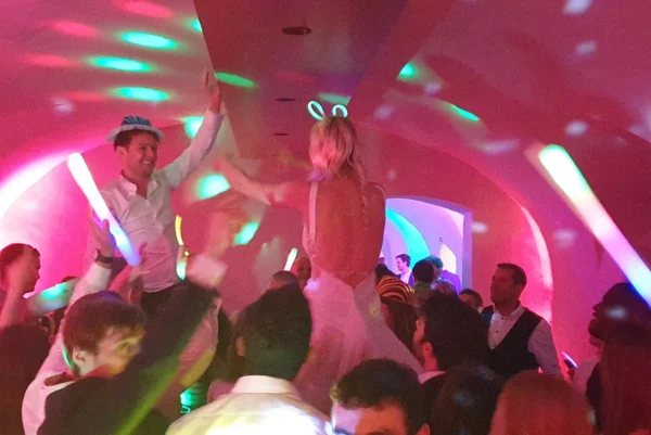 A bride and groom are lifted on chairs during a lively wedding celebration, surrounded by guests dancing under colourful lights in a festive, arched room.