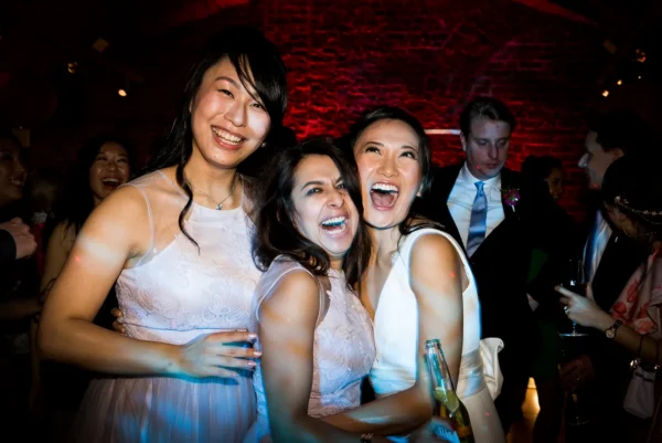 Three women in formal dresses smile and laugh together at a lively party, embracing each other. Other guests, including a man in a suit, are visible in the background under colourful lighting from a wedding DJ London.
