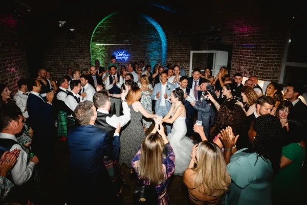 A bride and groom dance to their London wedding DJ closely surrounded by cheering wedding guests at RSA House with colourful neon and spot lighting. Many people clap and smile, creating a joyful, celebratory atmosphere.