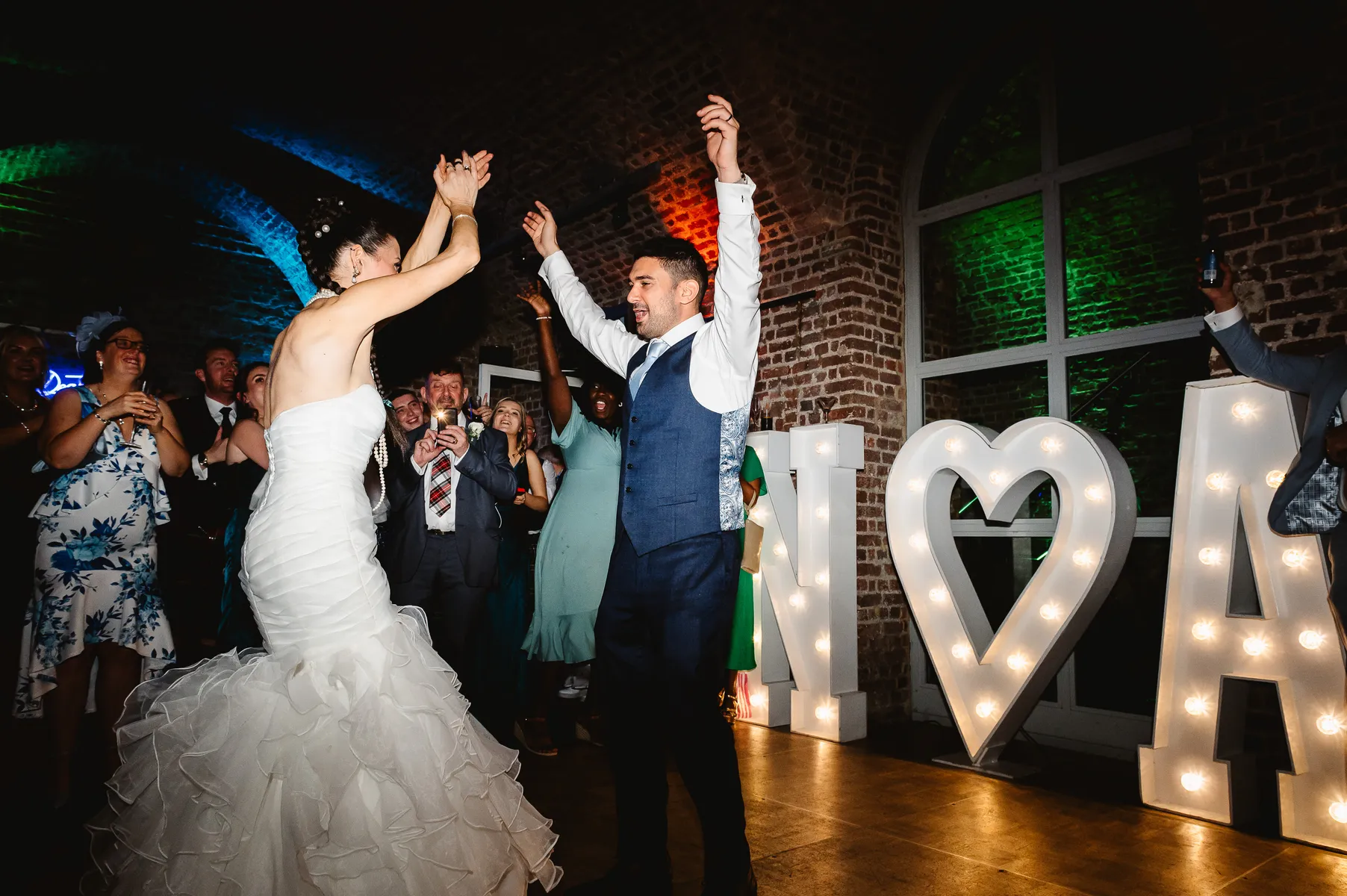 A bride and groom dance joyfully at their wedding reception at their wedding at RSA House, London, surrounded by cheering guests. Large illuminated letters and a heart shape glow in the background as the wedding DJ keeps the celebration lively against a brick wall.