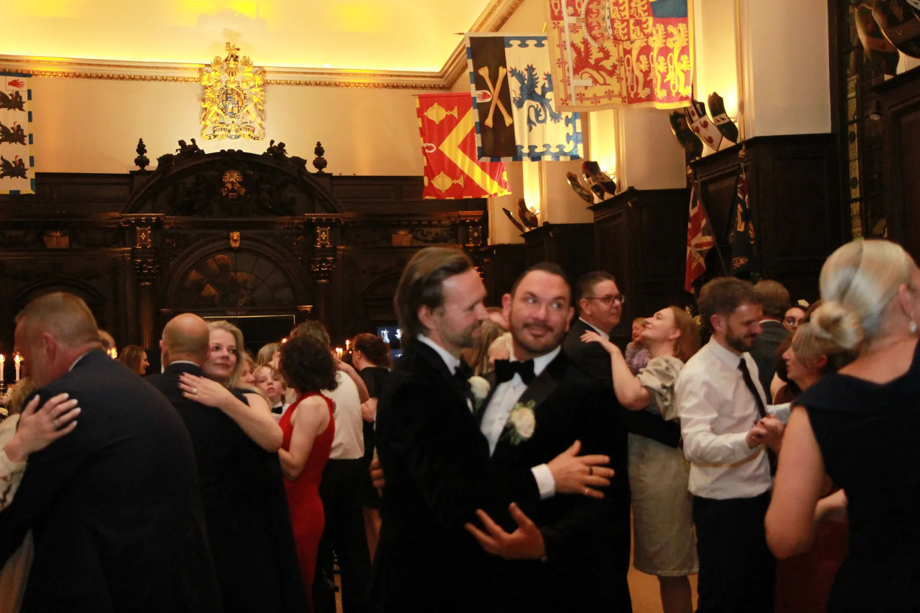 Two bridegrooms in dinner jackets do their first dance together at their wedding at Stationers Hall in London, which is decorated with colourful heraldic banners, while a wedding DJ sets the mood. The warmly lit room is filled with couples and guests in formal attire enjoying the celebration.