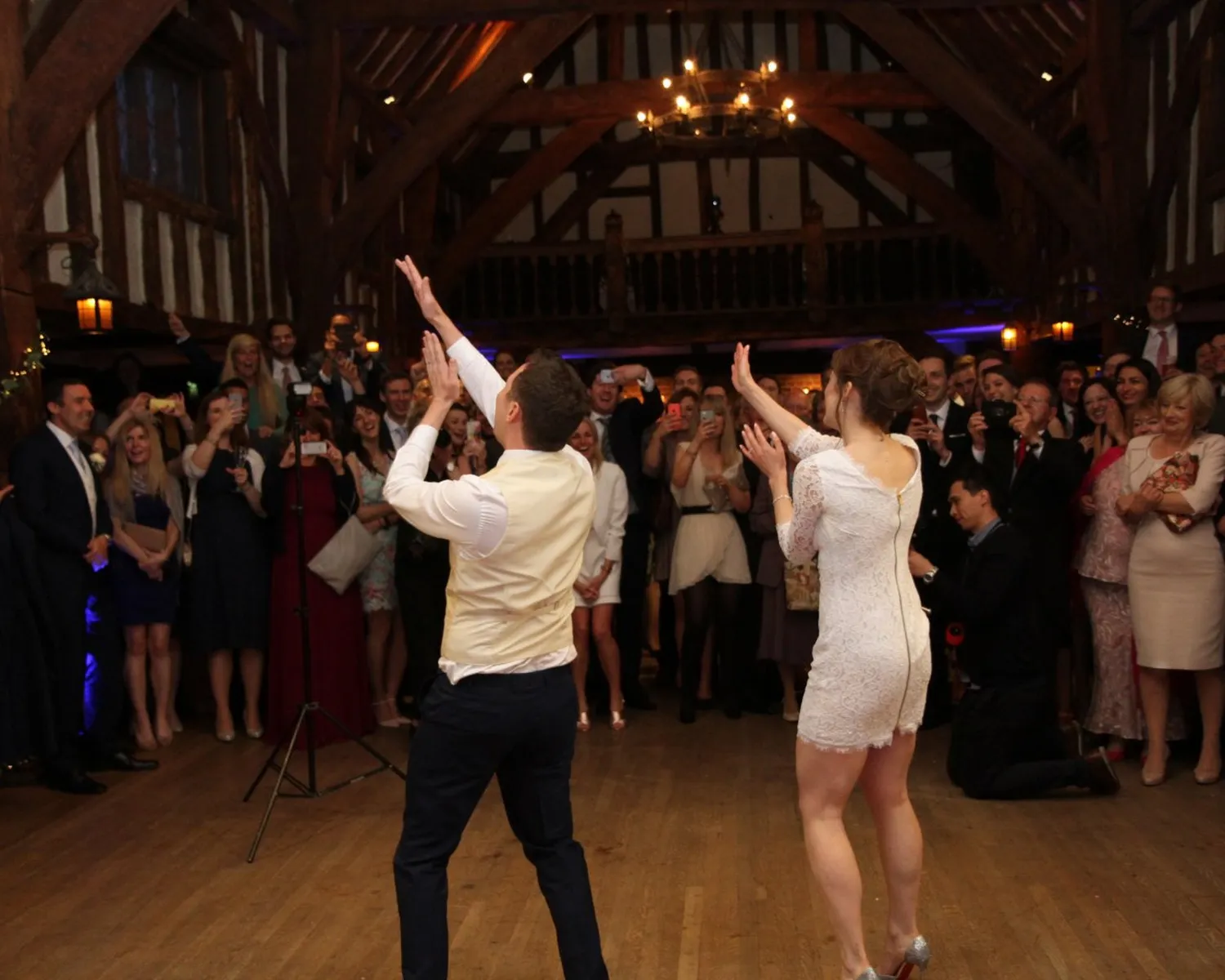 A couple dances energetically in the centre of a wooden hall, surrounded by cheering and smiling guests. The audience watches, some taking photos, under warm lighting and exposed wooden beams.