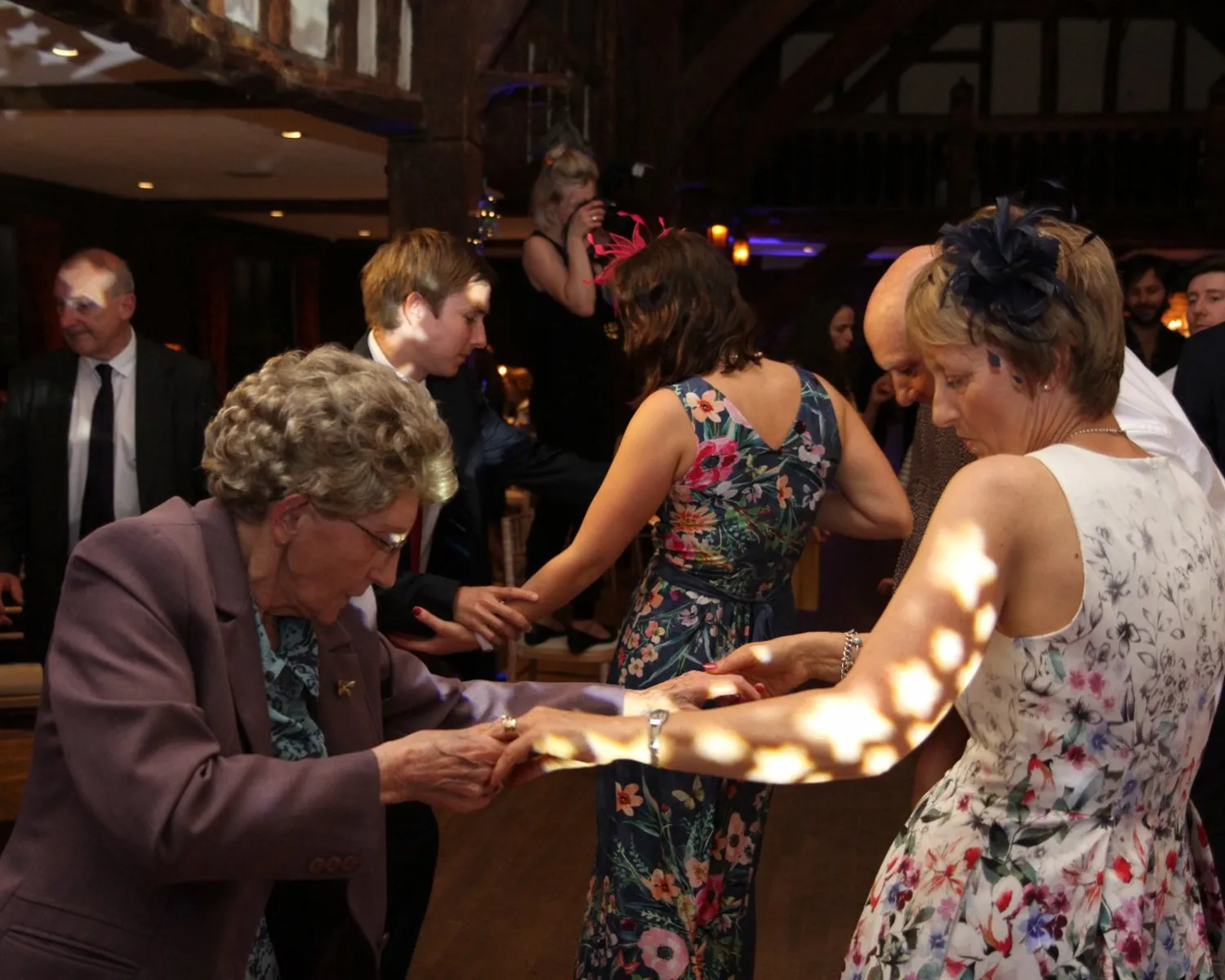 A group of people of various ages dance together at an indoor event, dressed in formal and floral attire under warm lighting, with a wooden-beamed ceiling visible above.