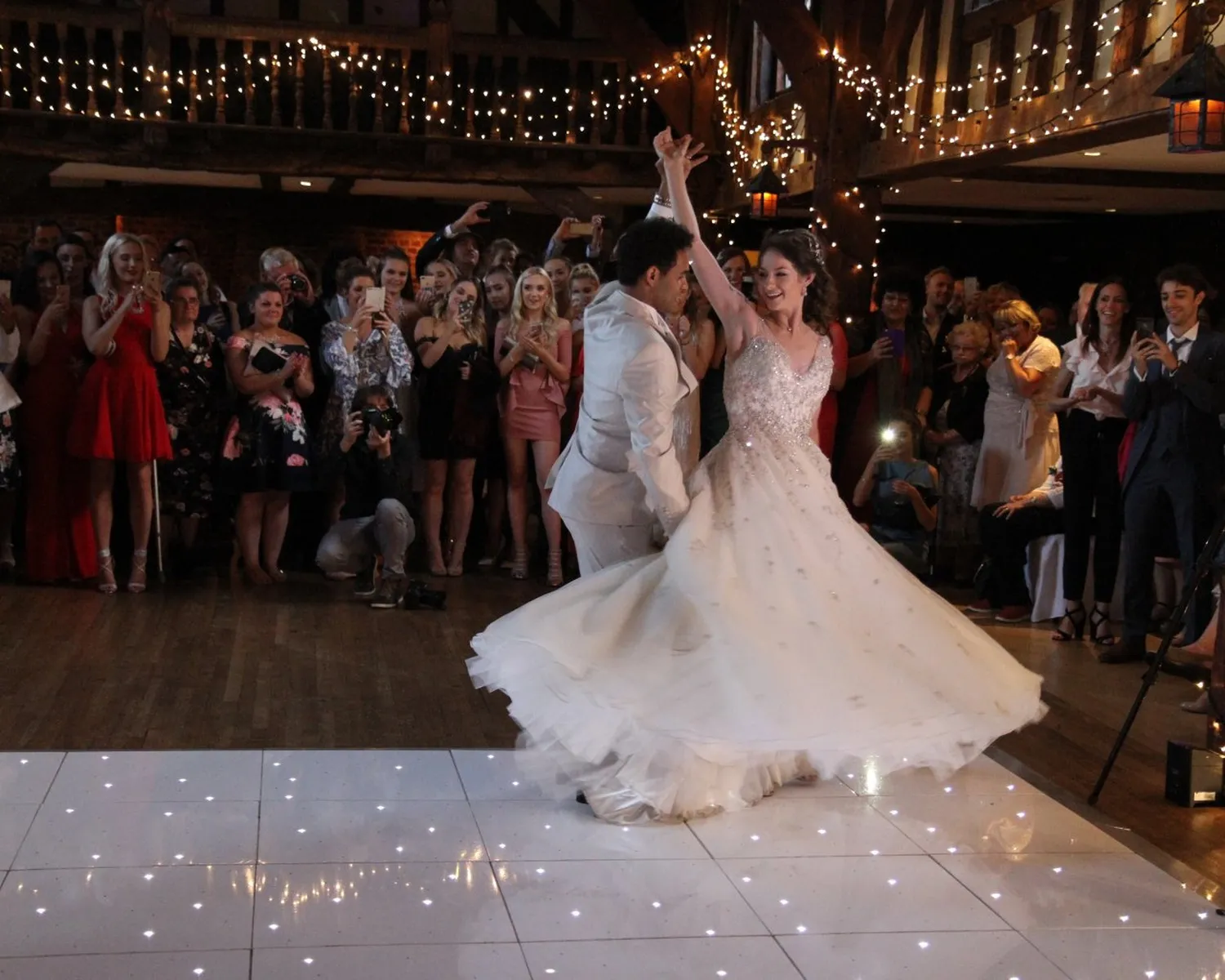 A bride and groom dance together on a lit-up floor as guests watch and celebrate at a wedding reception decorated with fairy lights. The bride's dress swirls as they spin.