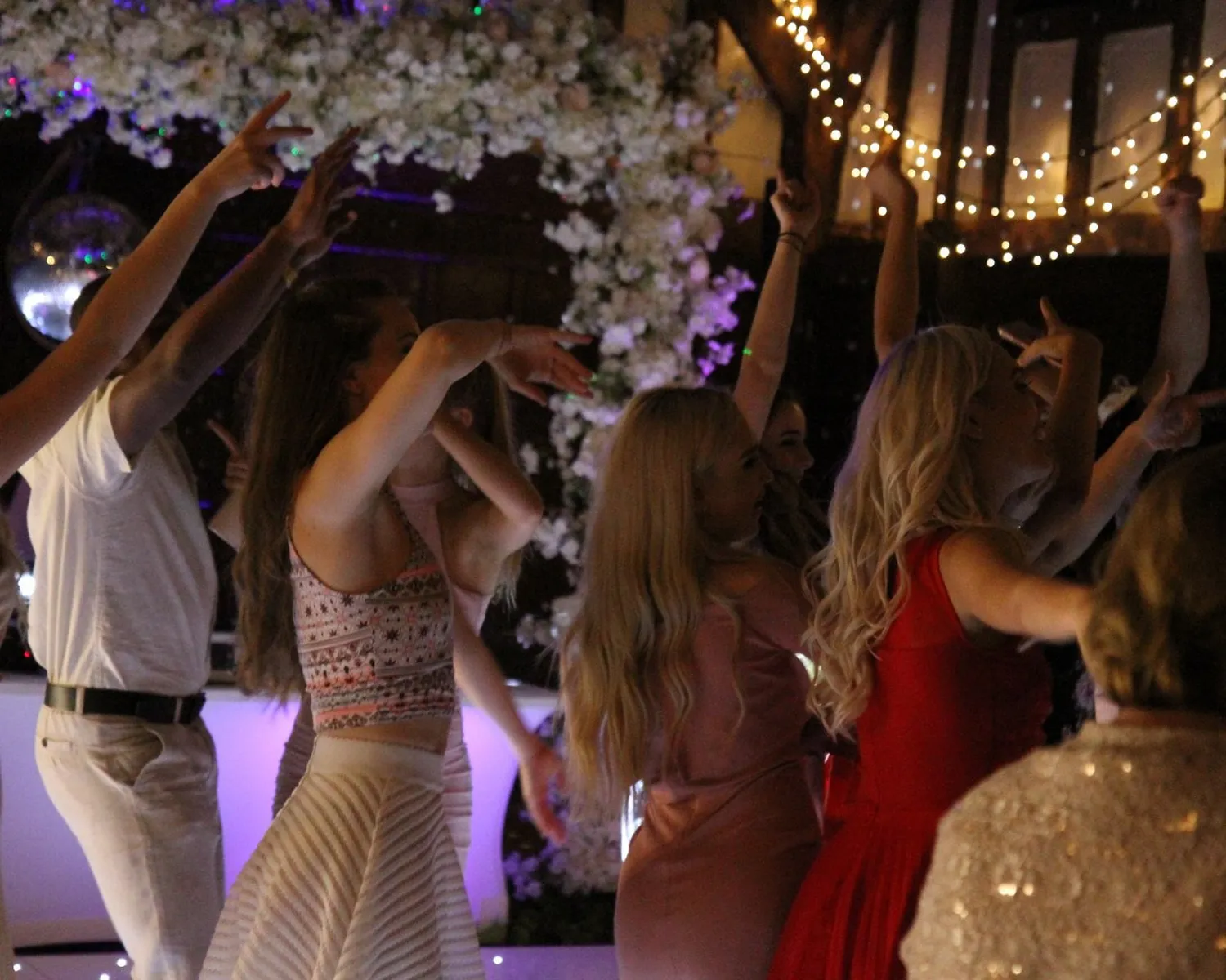A group of people dressed in semi-formal attire dance together at a lively indoor party, surrounded by floral decorations and fairy lights.