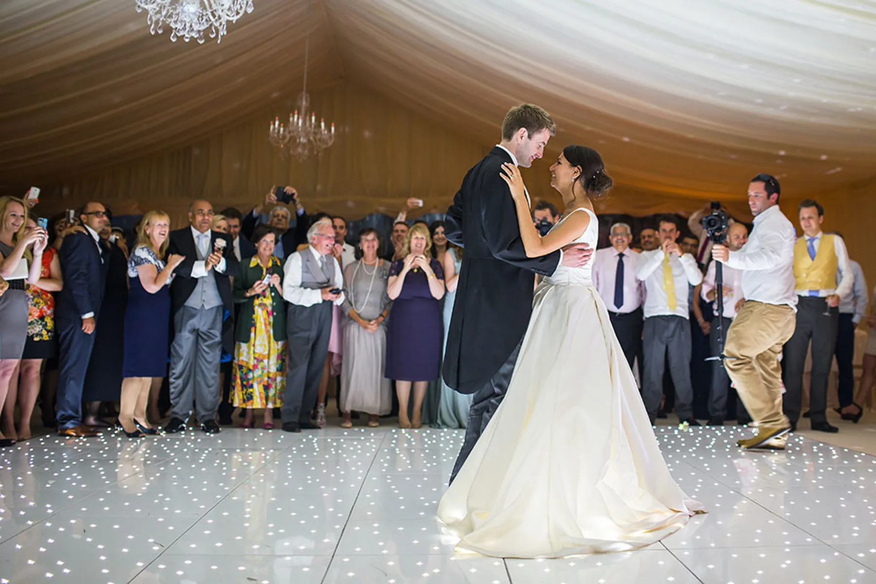 A bride and groom share their first dance on a sparkling white floor in a decorated marquee, surrounded by smiling guests who watch and take photos.