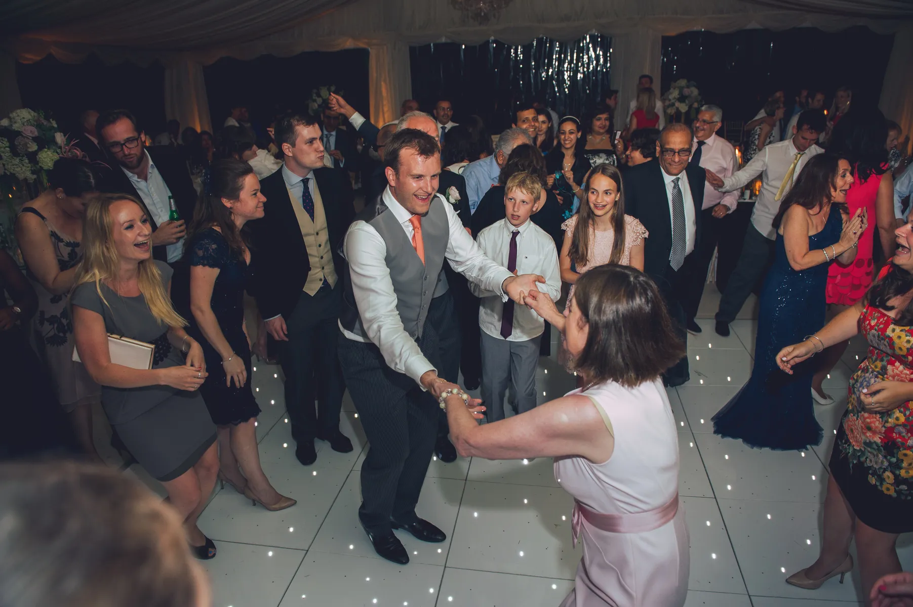 A joyful group of people dance together at an indoor event, with a man in a waistcoat and tie smiling as he holds hands and dances with a woman in a light pink dress; others watch and celebrate around them.