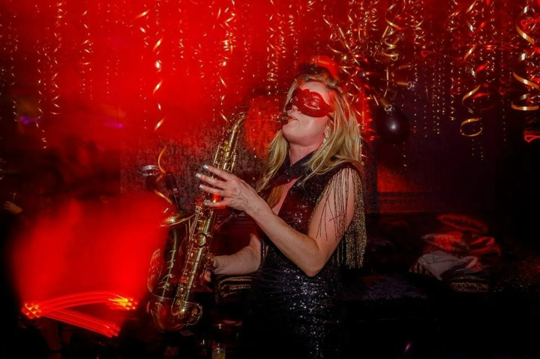 A woman in a red mask and black dress plays a saxophone alonside the DJ at a party, surrounded by red lighting and gold streamers hanging from the ceiling.