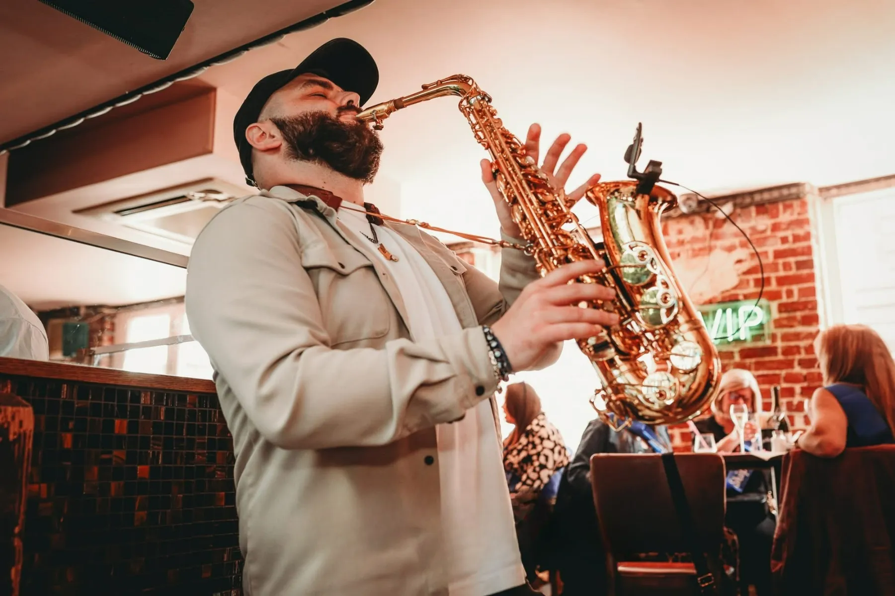 A man wearing a cap and light-coloured shirt plays a saxophone passionately indoors alongside a DJ as part of a DJ Live experience, with people sitting at tables and a neon sign visible in the background.