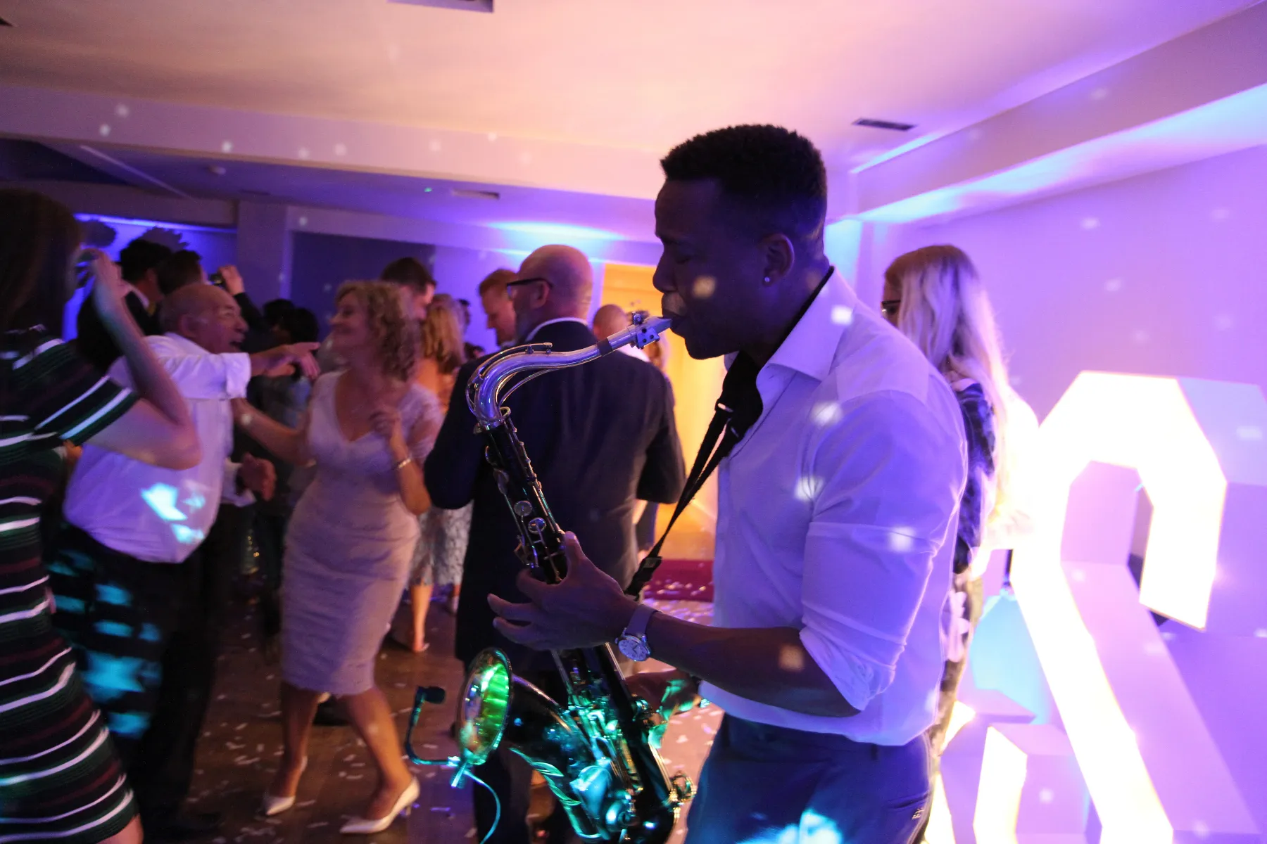 A man plays a saxophone alongside a wedding DJ for a DJ live performance at a lively indoor reception party, surrounded by people dancing under colourful lighting, with disco light spots scattered across the room.