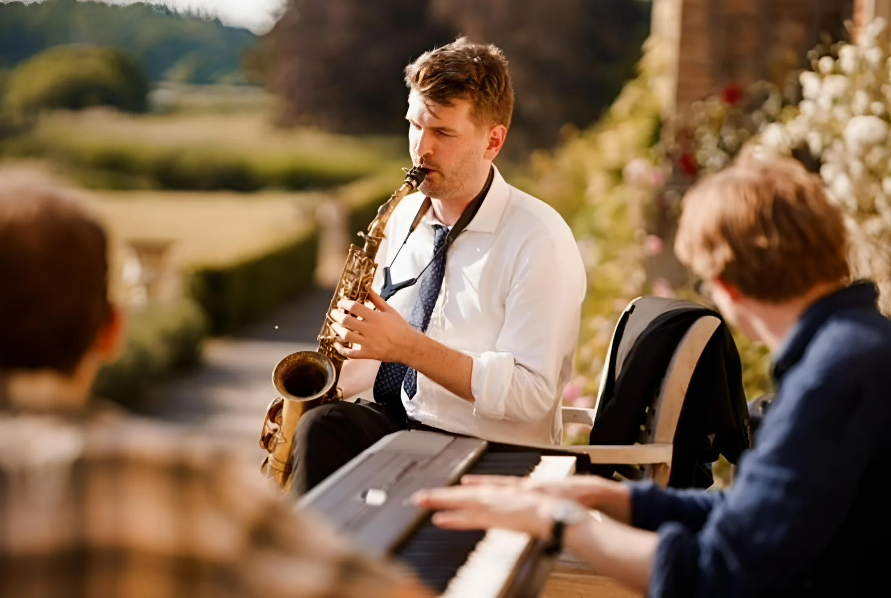 A man in a white shirt plays a saxophone outdoors while another person, seen from behind, plays a keyboard. They are surrounded by greenery and sunlight, suggesting a relaxed, outdoor music session.