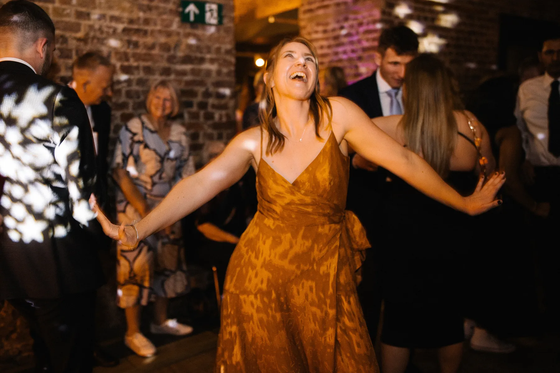 A woman in a patterned orange dress joyfully dances at a party, arms outstretched, smiling and looking up. Several people in formal attire stand and watch in the softly lit room with exposed brick walls.