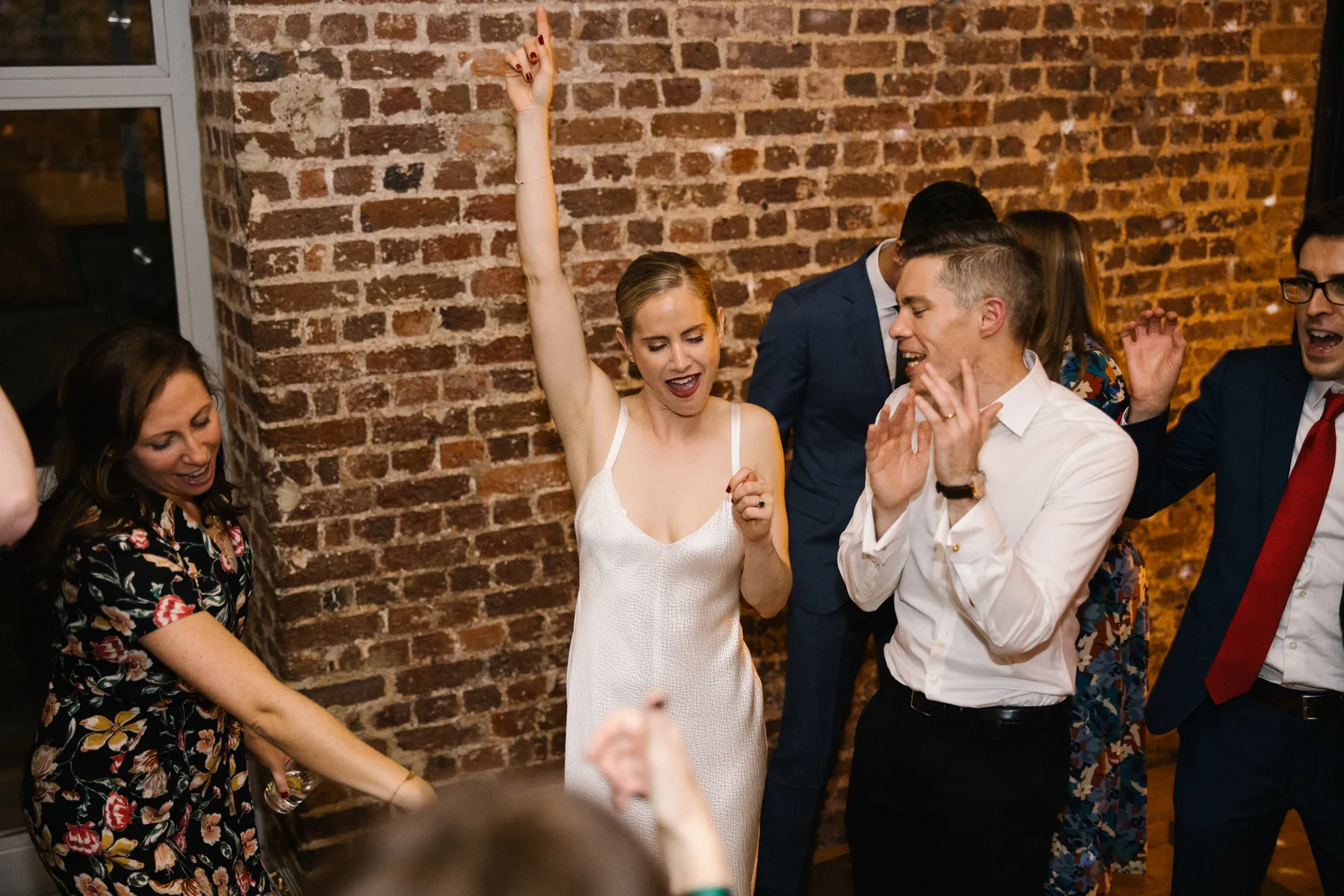 A group of people are dancing and celebrating indoors near a brick wall. A woman in a white dress raises one arm and smiles, whilst others around her clap and move to the music.