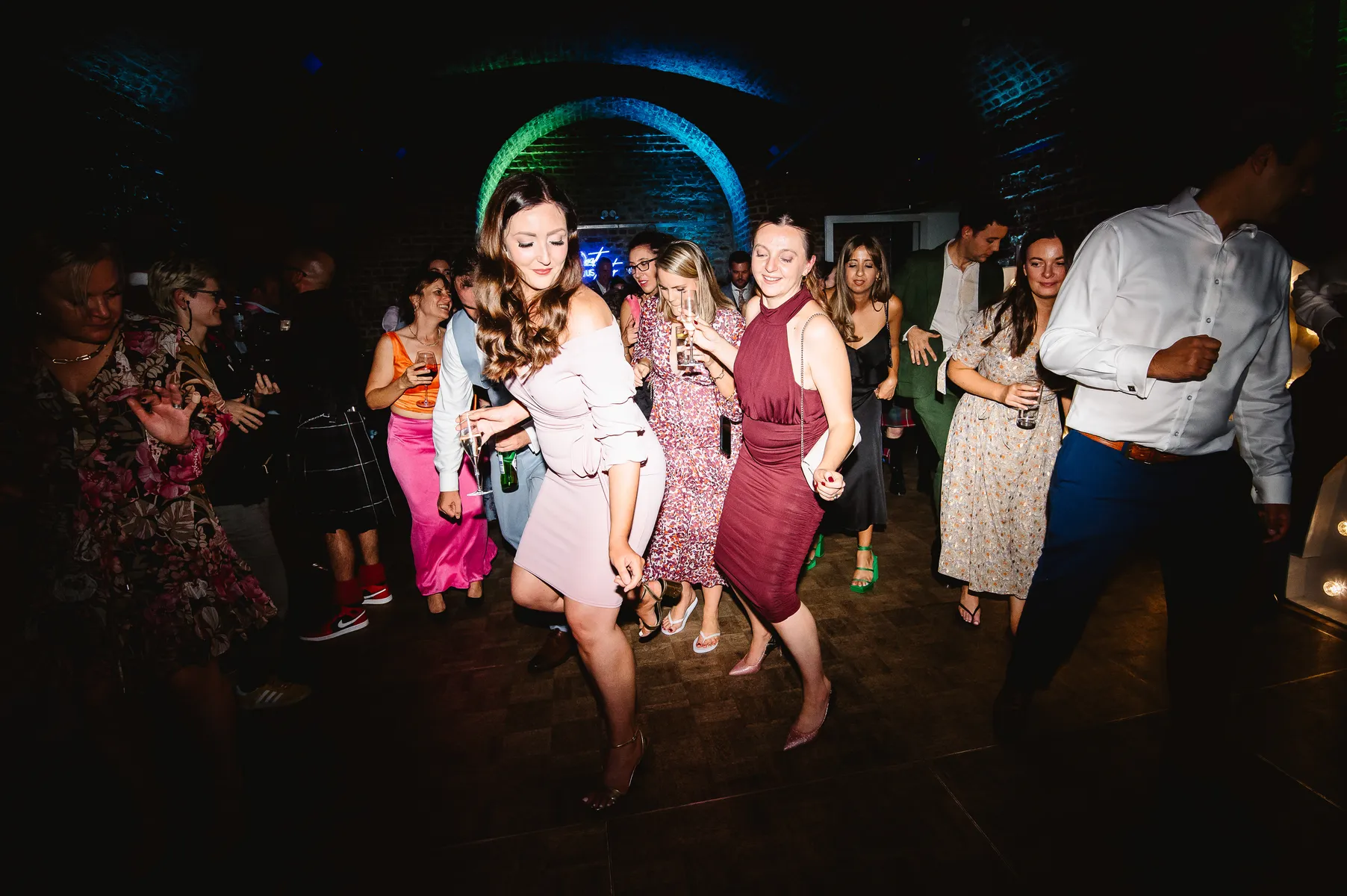 A group of people, mostly women in colourful dresses, dance energetically on a darkened dance floor under colourful lights at a lively indoor party at RSA House with a talented wedding DJ.