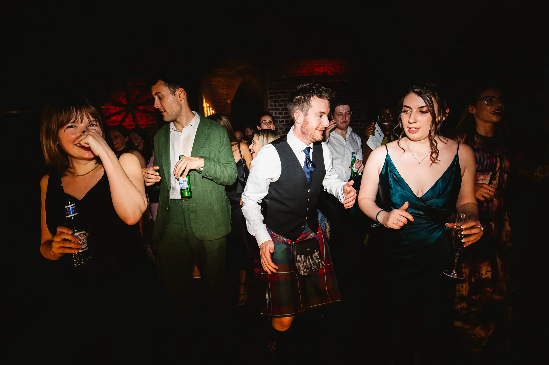 A group of people dancing and laughing at a party, with a wedding DJ setting the mood at RSA House; one person in a green suit, another in a kilt and waistcoat, holding drinks amid dim lighting and a festive atmosphere.