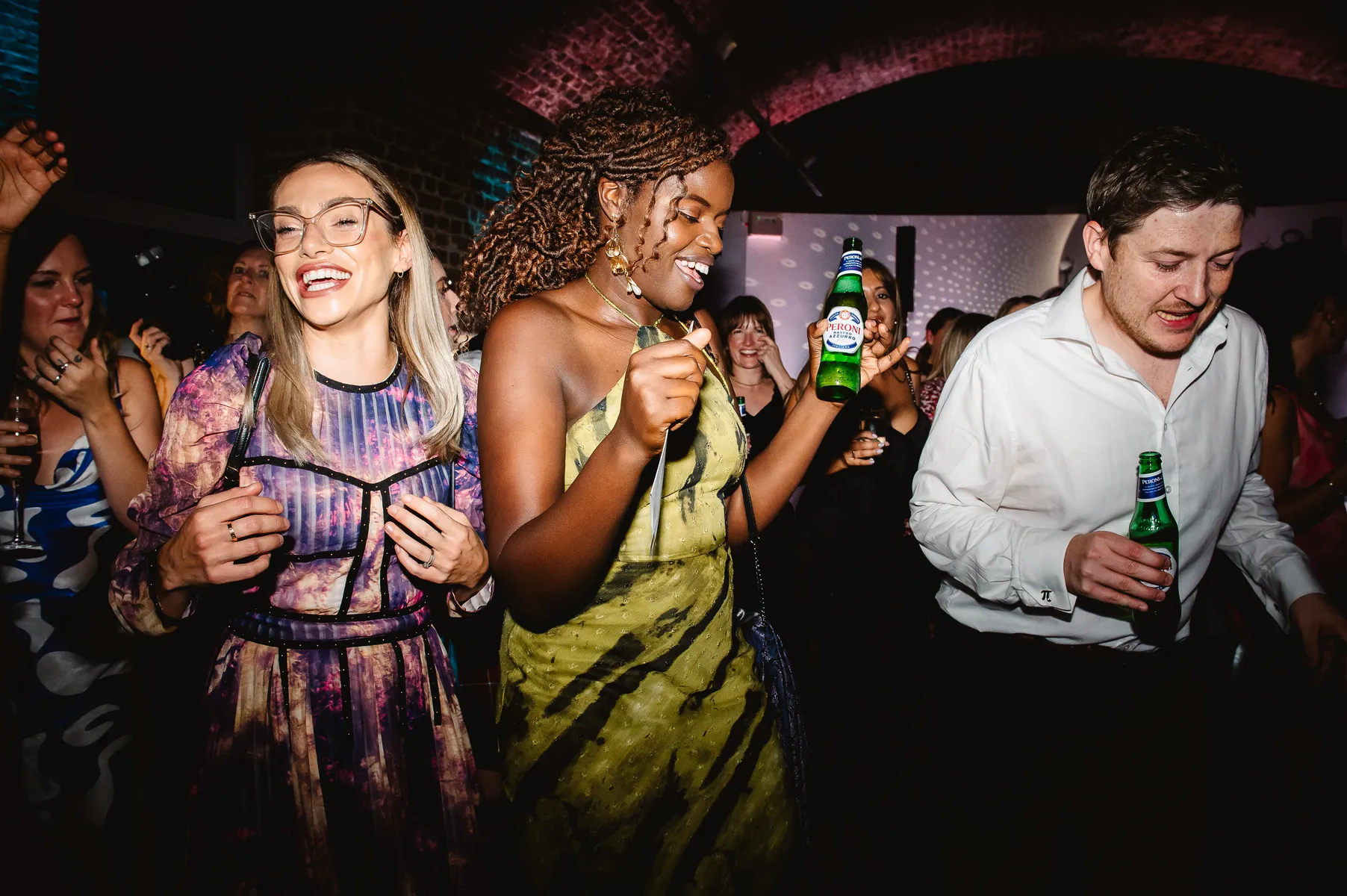Three people dance and smile at a lively party, holding drinks. The crowd behind them enjoys the celebration under colourful lights in the dimly lit RSA House, as a wedding DJ keeps the energy high.