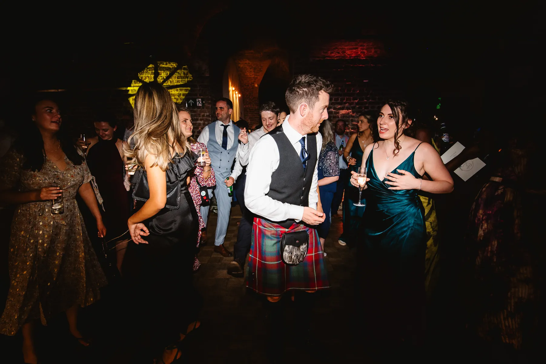 A man in a kilt and a woman in a teal dress are talking and smiling on a crowded dance floor at RSA House, with the wedding DJ keeping the party lively amid guests in formal and colourful outfits.