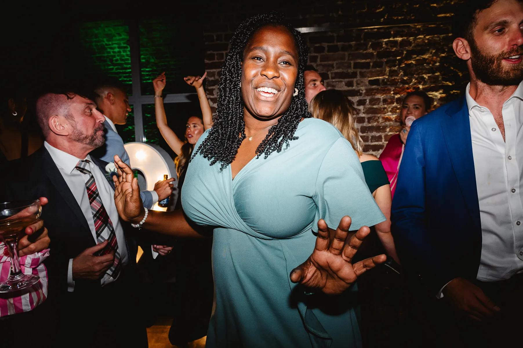A woman in a light blue dress smiles and dances at a lively indoor party, surrounded by people in formal attire, with brick walls and colourful lights in the background.