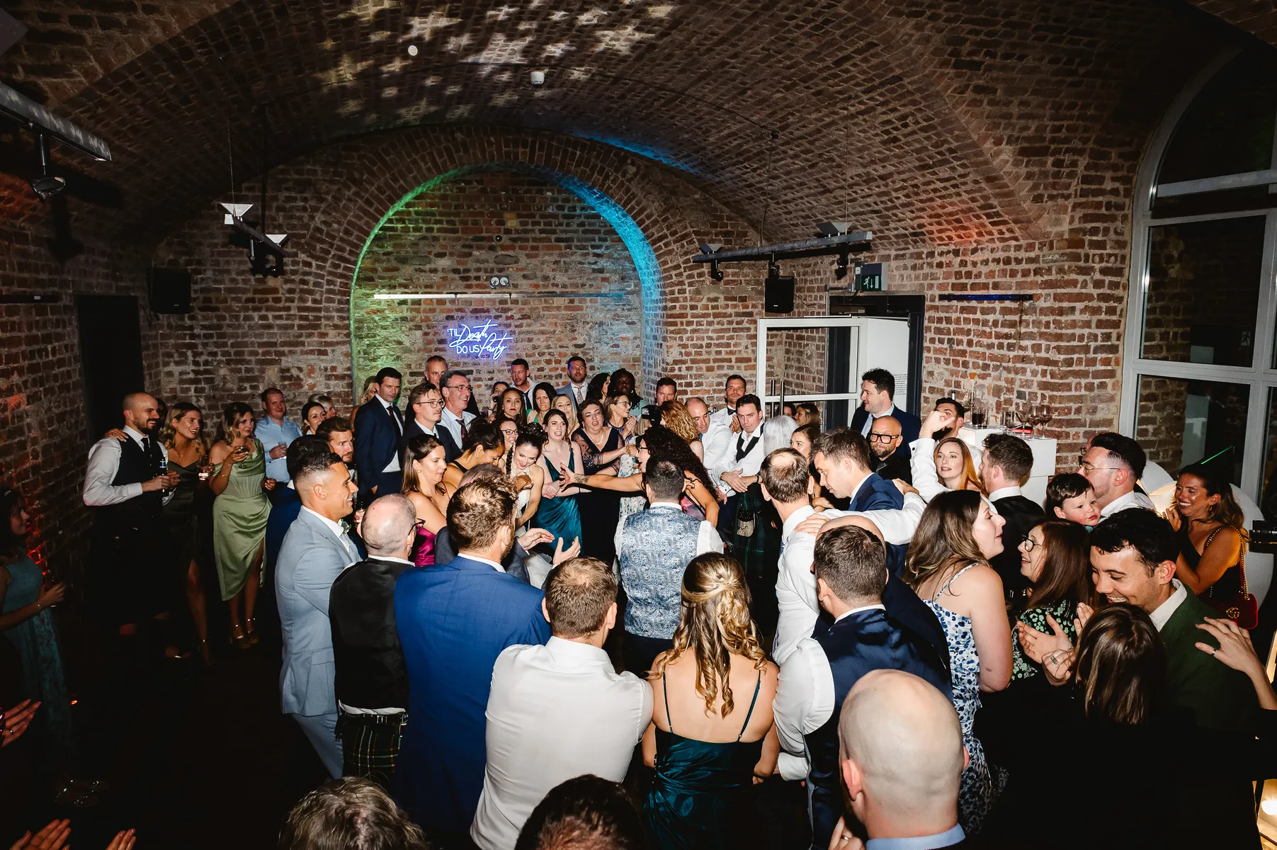 A large group of people dressed formally are gathered closely together, dancing and celebrating under arched brick ceilings at a lively indoor event, possibly a wedding reception. Colourful lights illuminate the scene.