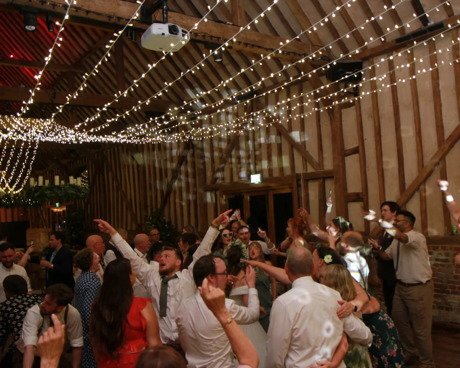 A lively wedding reception in a rustic barn, illuminated by fairy lights and energised by a talented wedding DJ Northamptonshire. Guests dance closely together, arms raised in celebration, fully immersed in the festive atmosphere.