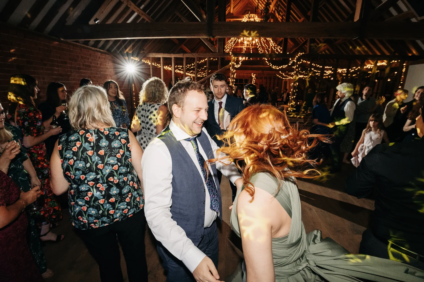 A man in a suit and a woman with red hair in a green dress dance energetically under fairy lights at a lively indoor wedding reception, as the wedding DJ Northamptonshire keeps guests smiling and the dance floor buzzing.
