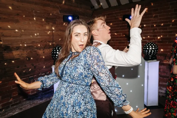 A woman in a blue floral dress and a man in formal wear stand back to back, striking playful dance poses at a party with disco lights, courtesy of a wedding DJ Northamptonshire, and a wooden and brick wall in the background.