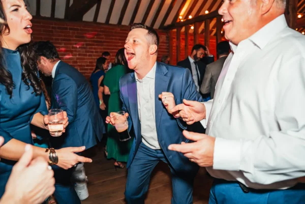 A group of people in formal attire enthusiastically dancing at an indoor event, smiling and holding drinks, with warm lighting and wooden beams in the background—brought to life by a top Wedding DJ Northamptonshire.