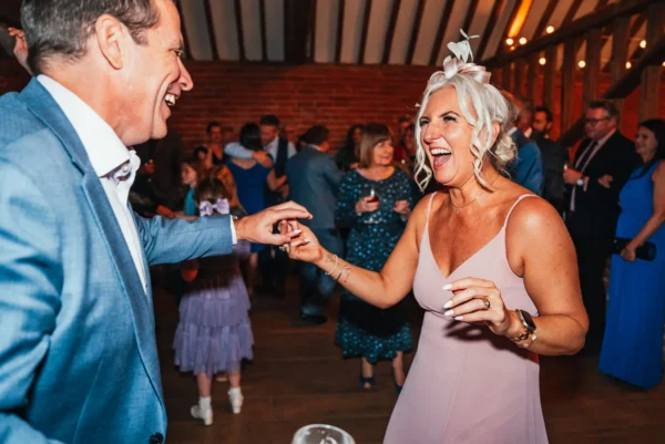 A smiling man and woman in formal attire laugh and dance together at a lively indoor wedding, with other guests mingling in the background, as the wedding DJ Northamptonshire keeps the energy high.