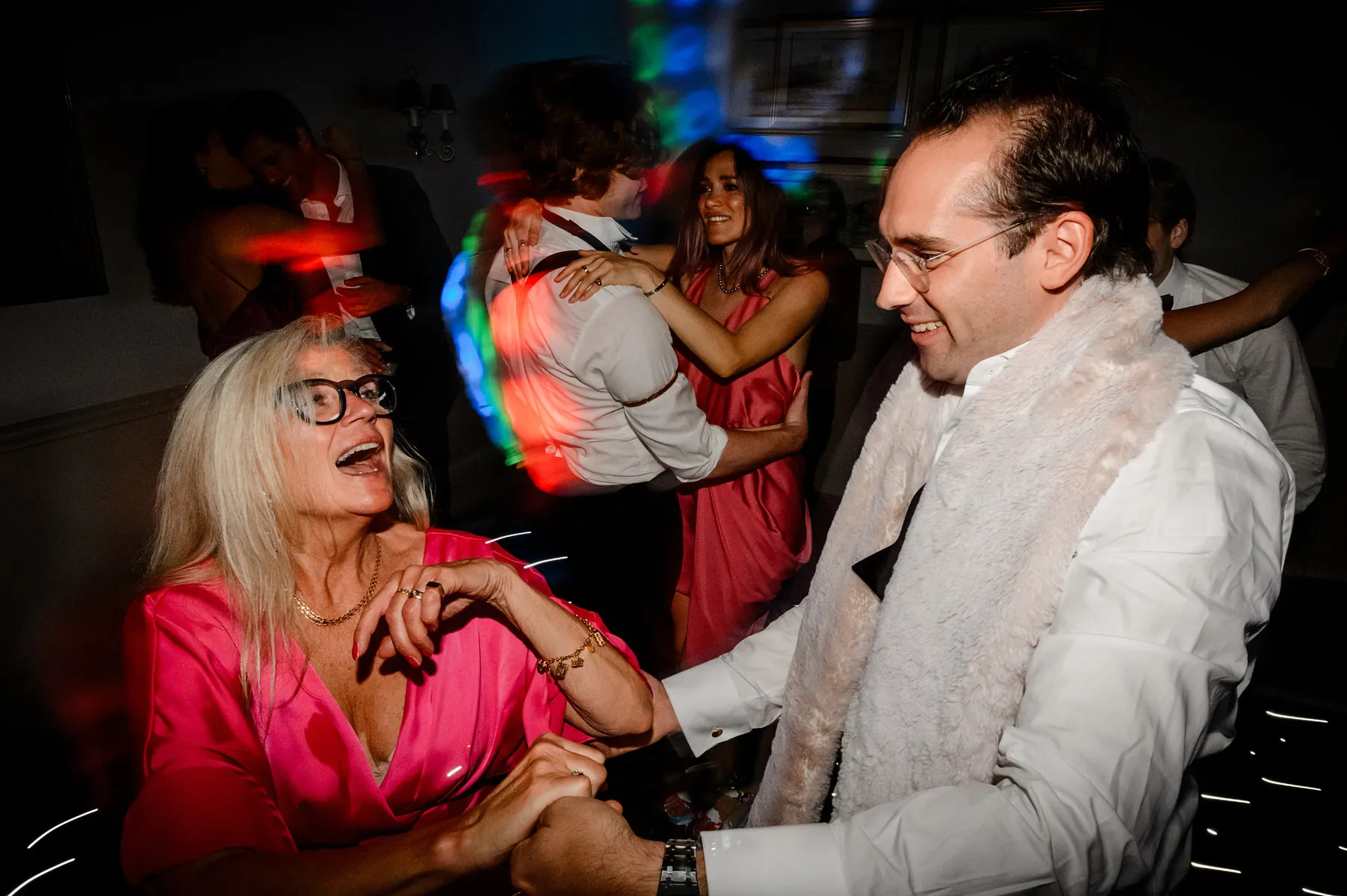 A joyful older woman in a bright pink dress and glasses dances with a man in a suit wearing a white feather boa at a lively party, as the wedding DJ Oxfordshire keeps guests moving on the colourful, dimly lit dance floor.