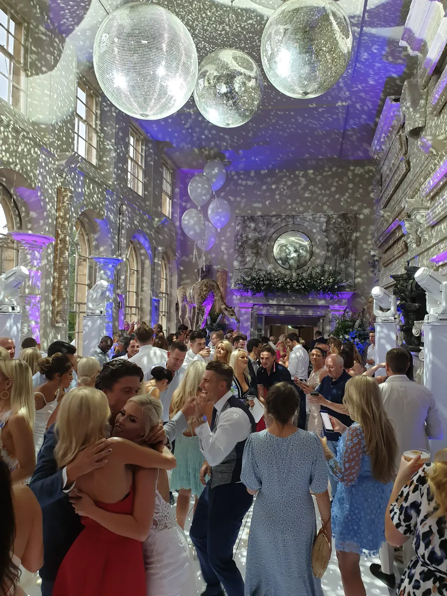 A lively wedding reception with guests dancing and mingling in a grand hall, decorated with disco balls, balloons, and purple lighting—expertly energised by a top wedding DJ in Oxfordshire as patterns of light shimmer across the walls and floor.