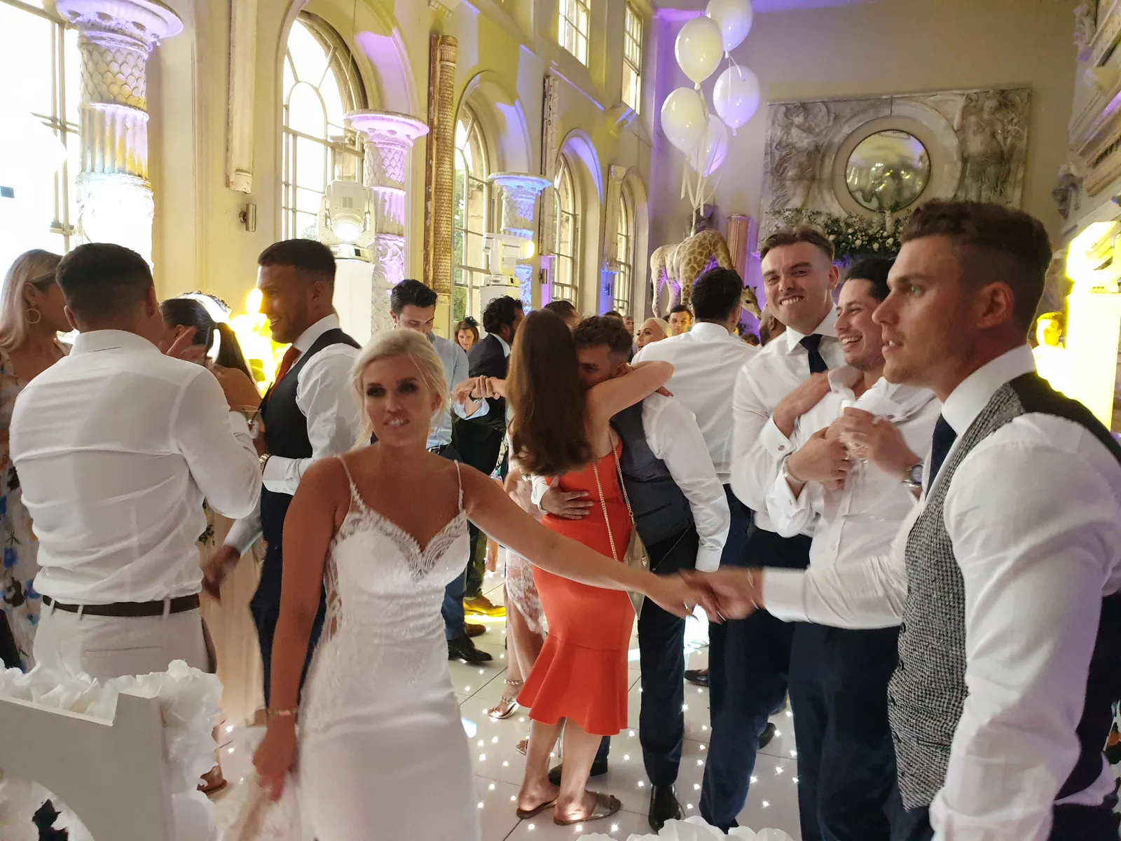 A bride in a white dress holds hands with a man on the dance floor at a lively Aynhoe Park wedding reception in Oxfordshire, as the DJ plays music. Guests in formal attire celebrate under tall windows and white balloons in the decorated hall.