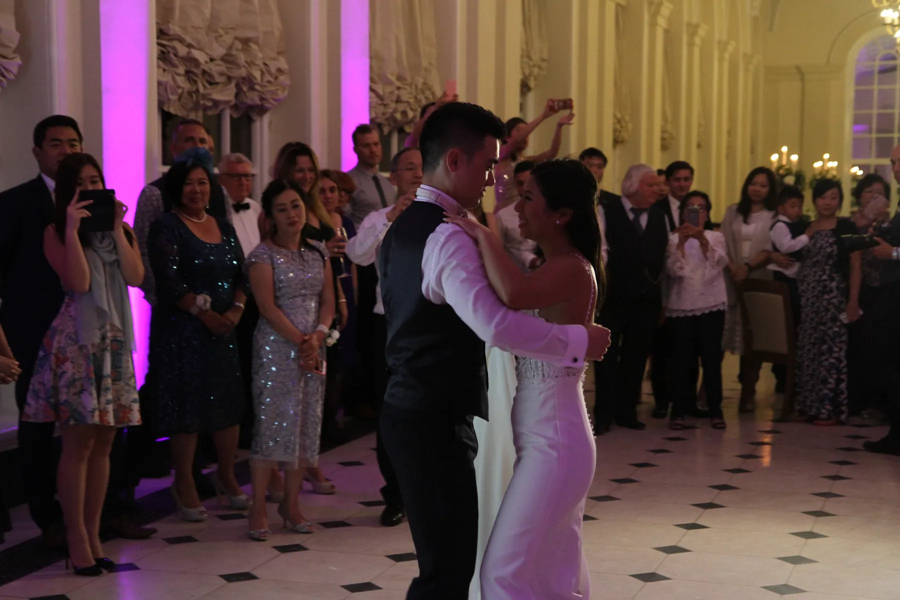 A bride and groom dance closely together at their wedding reception in Oxfordshire, surrounded by smiling guests taking photos in a grand, elegant hall with purple lighting, large windows, and music by a renowned wedding DJ at Blenheim Palace.