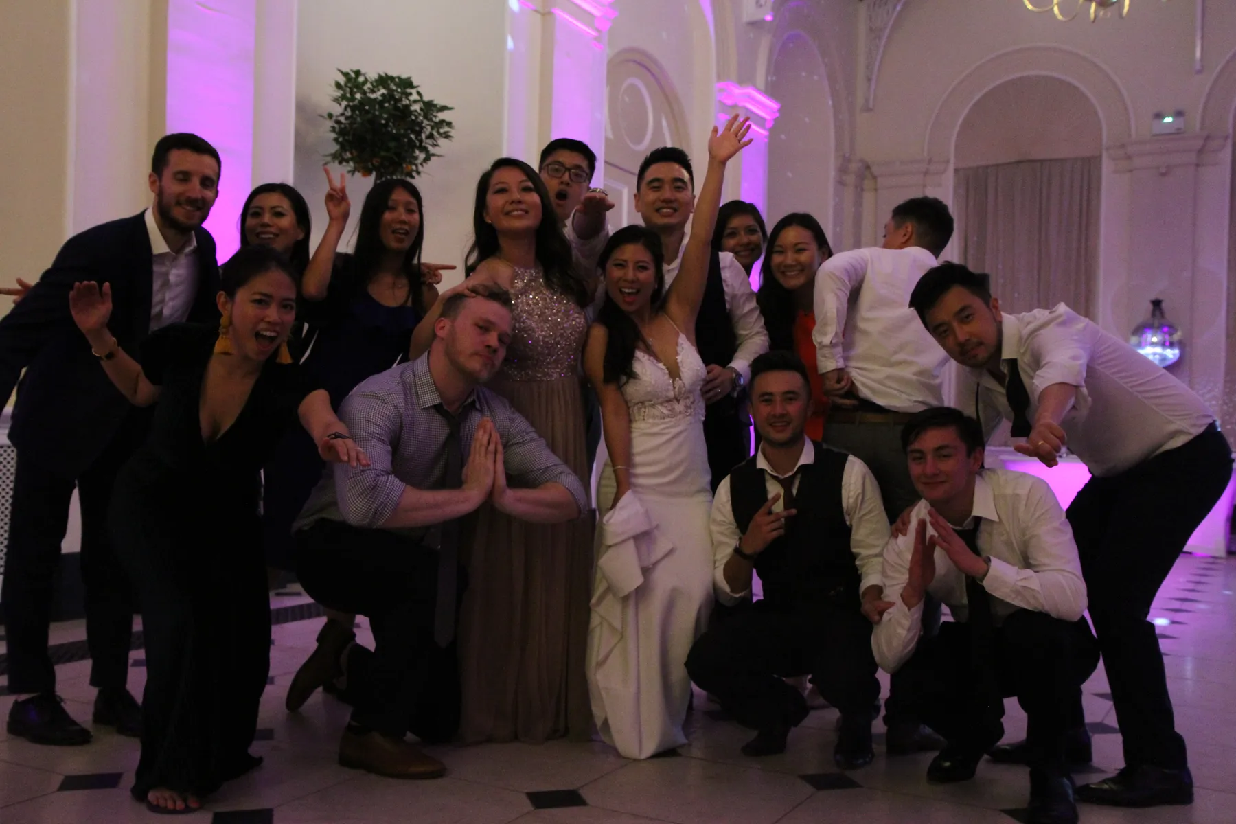 A group of people dressed formally, smiling and posing together at an indoor event with elegant white walls and purple lighting. The bride stands in the centre wearing a white gown, celebrating with friends as the wedding DJ Oxfordshire sets the mood.