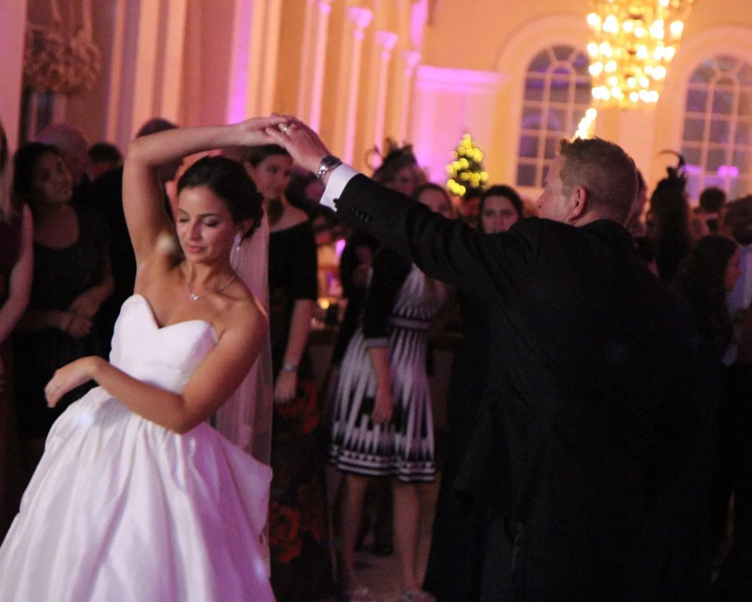 A bride in a white dress dances with a man in a suit at an elegant Oxfordshire event, surrounded by guests. Chandeliers and arched windows glow with pinkish light, as a wedding DJ at Blenheim Palace sets the perfect mood.