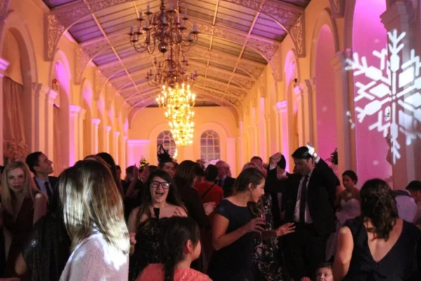 A group of people dressed in formal attire dance and socialise in a grand ballroom with chandeliers and arched windows. Pink lighting and a large snowflake projection decorate the festive scene.