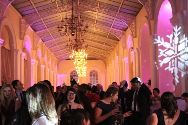 A group of people dressed in formal attire dance and socialise in a grand ballroom with chandeliers and arched windows. Pink lighting and a large snowflake projection decorate the festive scene.