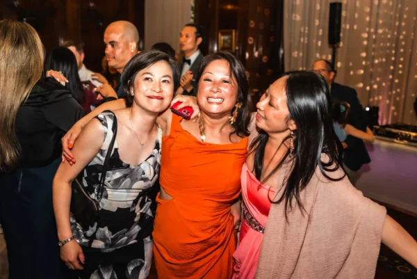 Three women smile and pose together at a lively indoor event, dressed in elegant dresses as guests mingle and dance to the tunes of a wedding DJ Oxfordshire. Warm lighting and festive decorations create a joyful atmosphere.