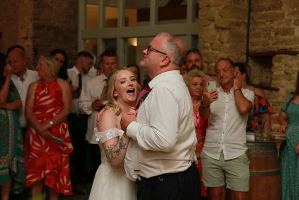 A bride and an older man, possibly her father, dance together at a wedding reception while guests watch and take photos in the background. The bride is smiling widely and wearing an off-shoulder dress.