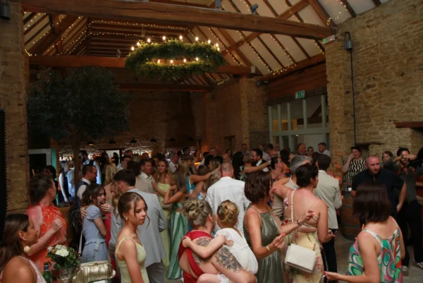 A large group of people dressed in formal attire gather inside a rustic barn-style venue with wooden beams and fairy lights, mingling and socialising at what appears to be a wedding in Oxfordshire with a wedding DJ at The Great Barn Aynho.