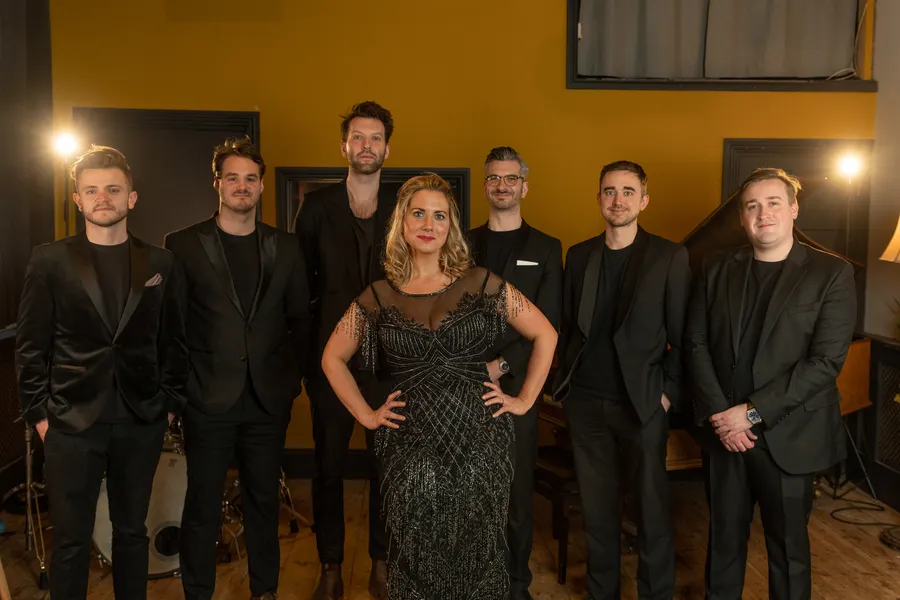 Seven people dressed in formal black outfits stand together indoors, with a jazz singer in an embellished dress at the centre and six men in suits around her, posing and smiling in a warmly lit room.