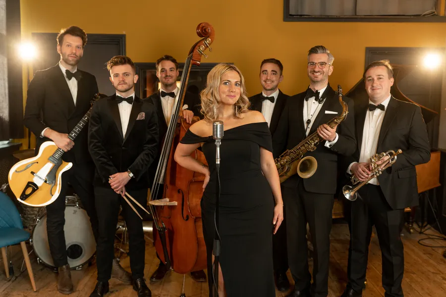 A jazz band of six men in dinner jackets with instruments and one jazz singer in a black dress stand together indoors; the woman stands at a vintage microphone, with guitars, a saxophone, trumpet, double bass, and drums visible.