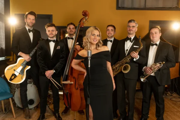 A jazz band of six men in dinner jackets with instruments and one jazz singer in a black dress stand together, smiling, in a warmly lit room. The singer is at the front, standing at a vintage-style microphone.
