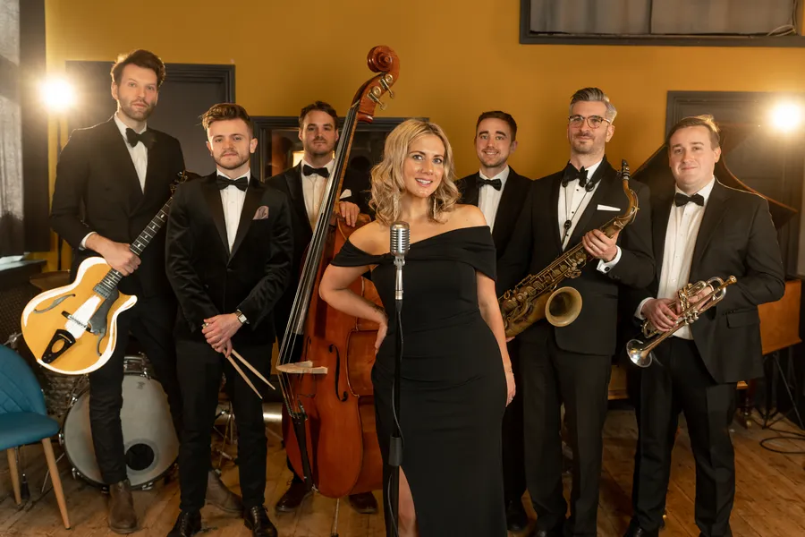A jazz band of six men in dinner jackets with instruments and one jazz singer in a black dress stand together, smiling, in a warmly lit room. The singer is at the front, standing at a vintage-style microphone.