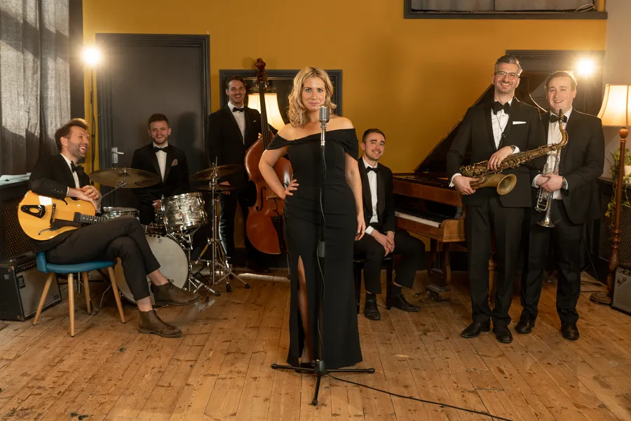 A jazz singer in a black dress stands at a microphone, surrounded by five men in dinner jackets holding musical instruments—guitar, drums, double bass, piano, and saxophone—in a warmly lit room with yellow walls.