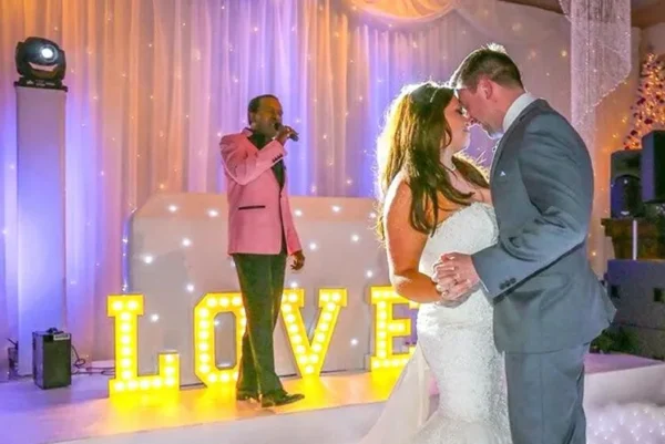 A bride and groom share their first dance at a wedding reception. Behind them, a singer in a pink jacket performs on stage, and large yellow marquee letters spell out LOVE, illuminated against a draped backdrop.