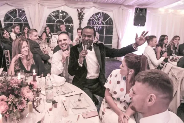 A lively man in a dark suit sings into a microphone among smiling guests at a formal event in a decorated marquee, with people seated at round tables enjoying the celebration.