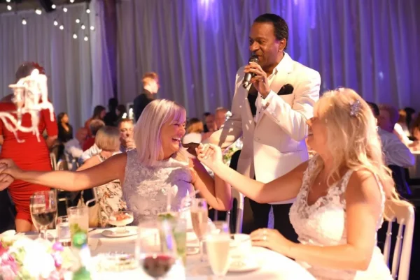 A man in a white suit sings into a microphone at an event, interacting with two smiling women in white dresses seated at a decorated table. Other guests and tables are visible in the softly lit background.