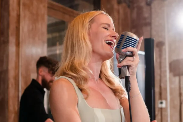 Wedding and events singer, Jenny, in a light dress sings passionately into a vintage microphone as a wedding singer indoors, with a blurred musician in the background and bright studio lights behind her.