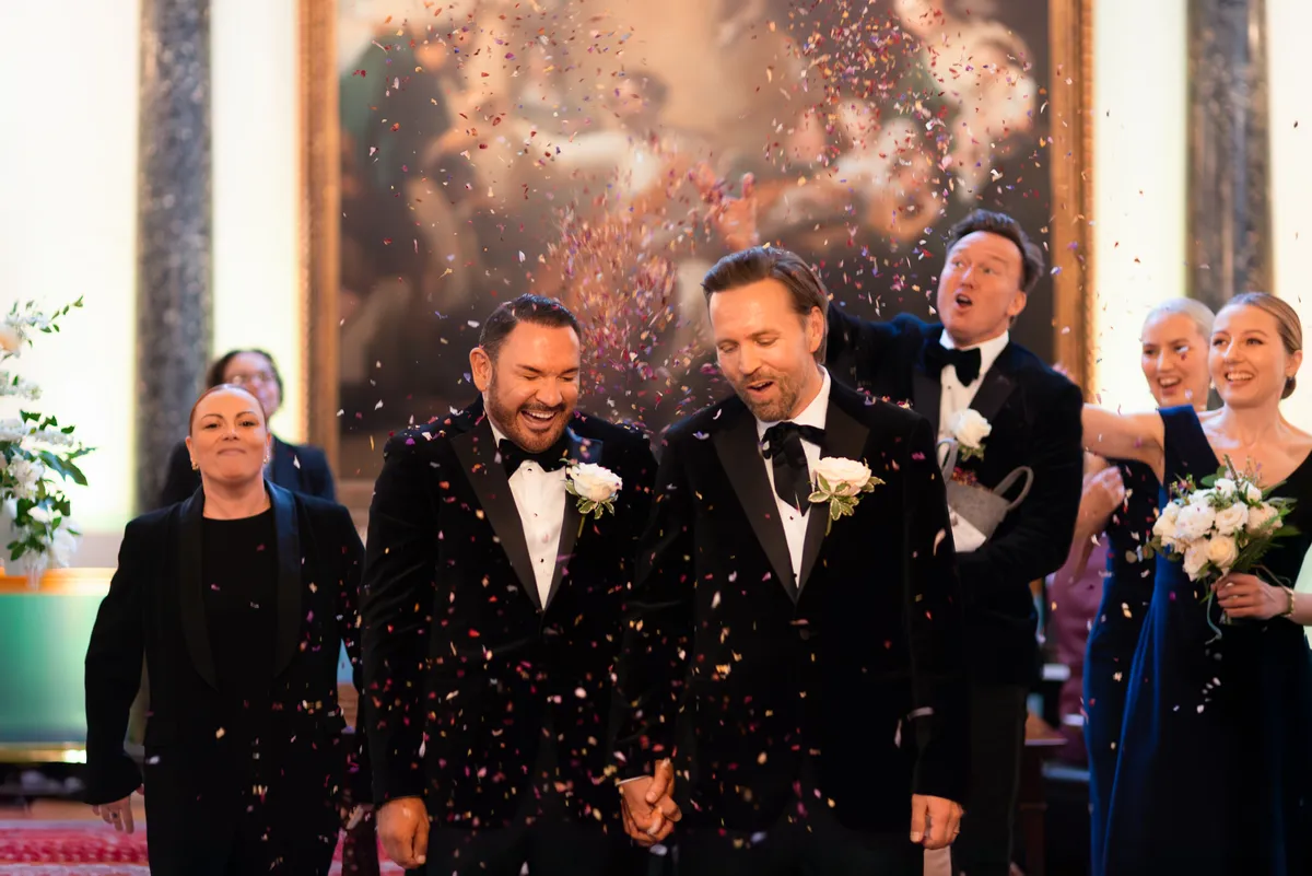 Two grooms in tuxedos walk hand in hand down the aisle, smiling joyfully as guests throw colourful confetti. People around them celebrate, holding bouquets and clapping, in a warmly lit indoor setting at a wedding at Stationers' Hall in London.