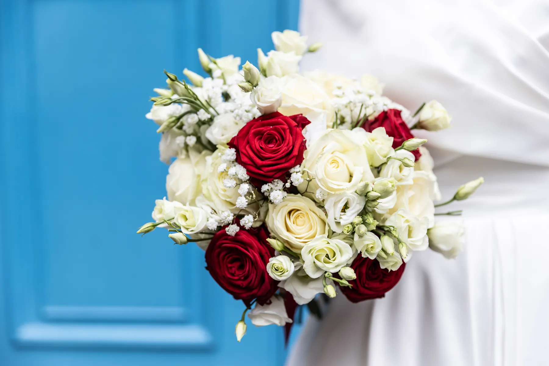 A person in a white dress holds a bouquet of red and white roses with small white flowers, standing in front of a bright blue background at Stationers' Hall.